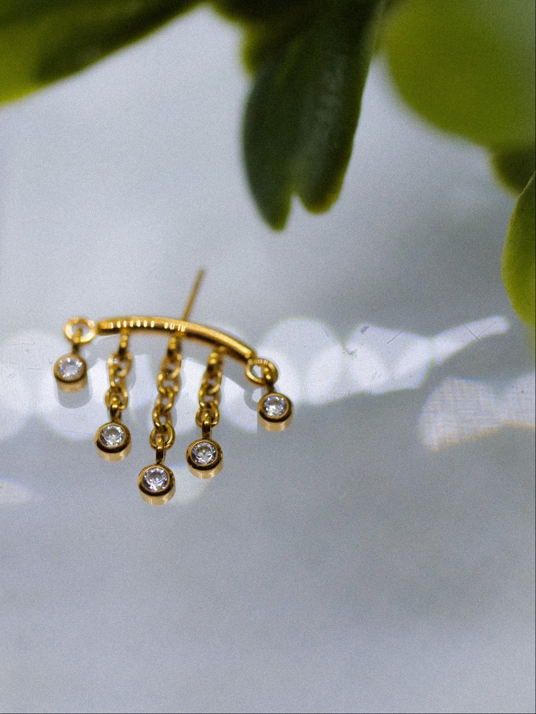 A gold crescent-shaped stud earring with five dangling chain segments, each ending in a small clear gemstone, photographed against a soft white background with blurred green leaves on the left.